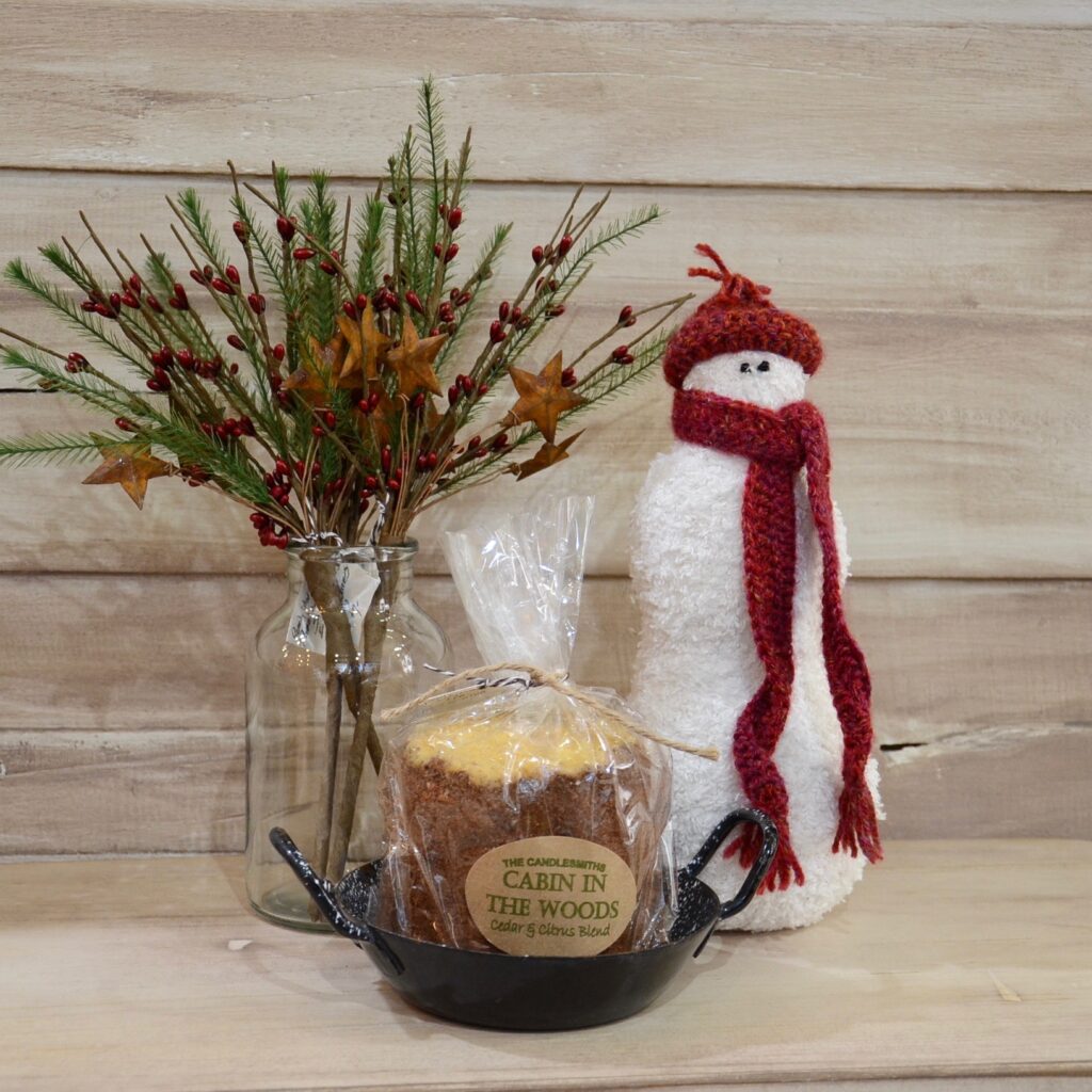 Staged holiday products including a glass jar with pine, berry and stars next to candle in tray and fabric snowman wearing a red hat and scarf.