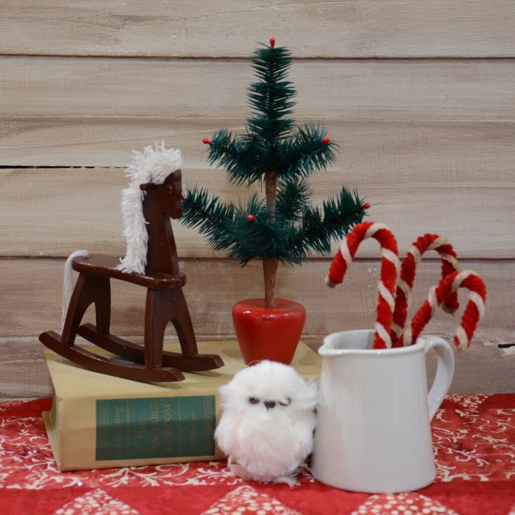 Holiday decor display with small wooden rocking horse, vintage book, feather owl, feather tree, and white ceramic pitcher with candy canes inside.